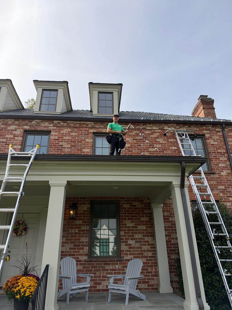 Green Zebra crew member on a porch roof installing C9 Christmas lights on a brick colonial